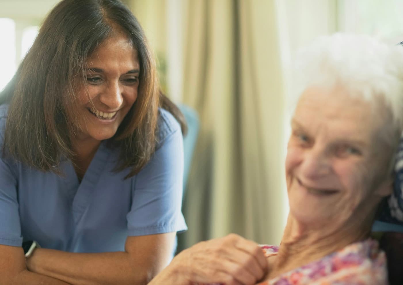 A community nurse and an older woman laughing warmly together during a home visit.
